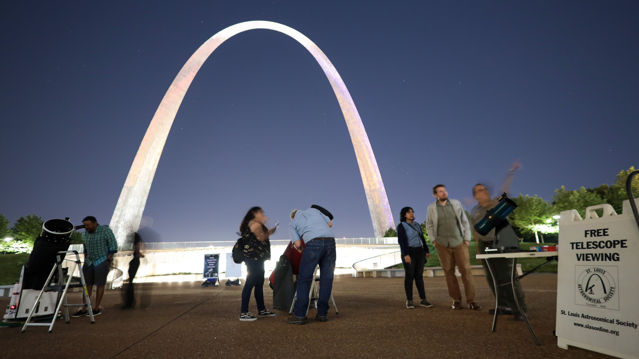 Visitors look into large telescopes sitting in front of the Gateway Arch during a Gateway to the Stars event