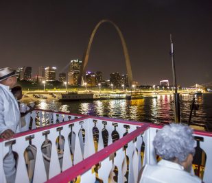 Guests sit on the top deck of the Riverboats at the Gateway Arch at night looking at the Gateway Arch illuminated