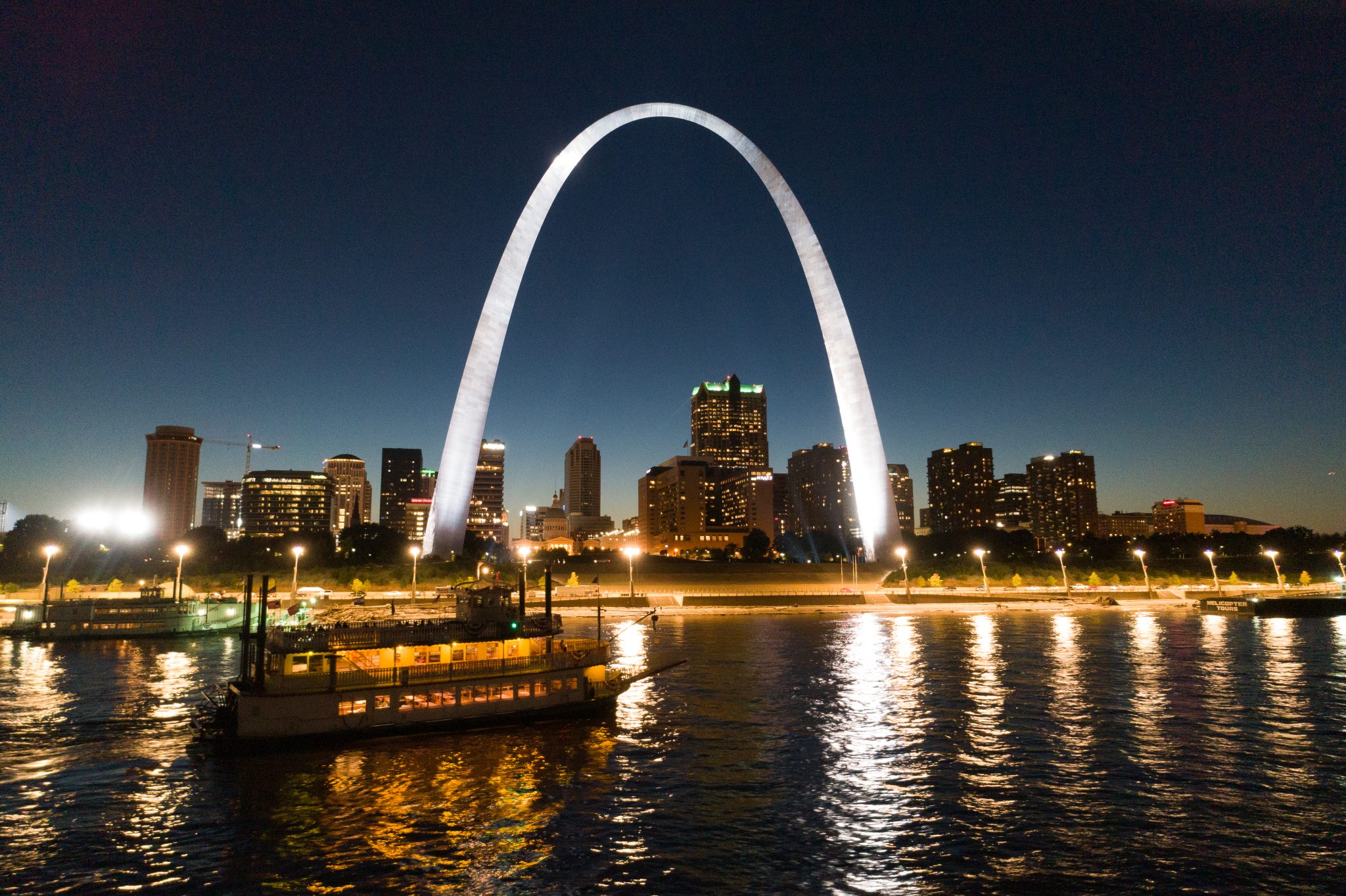 The Becky Thatcher riverboat cruises at night along the Mississippi River in front of the illuminated St. Louis Riverfront and Gateway Arch.