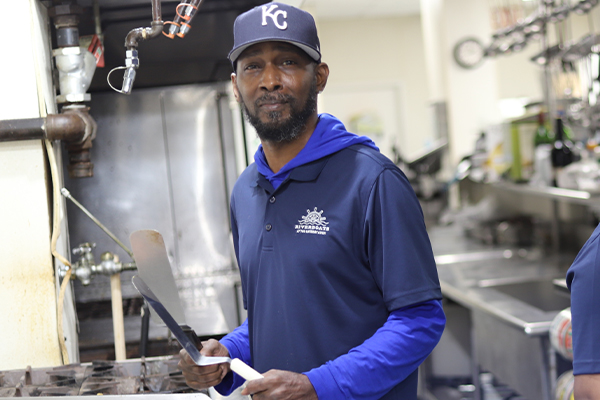 A chef smiles for a portrait while holding spatulas over a stovetop inside the galley at the Riverboats at the Gateway Arch.