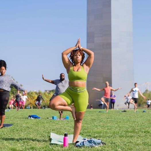 A woman stands on one leg in tree pose during a yoga session with dozens of other people under the Gateway Arch.