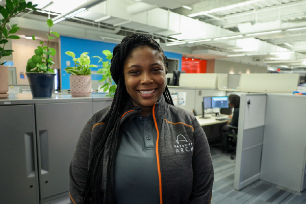 Female Call Center Agent smiles for a photo as she stands near cubicles decorated with plants and colorful artwork.